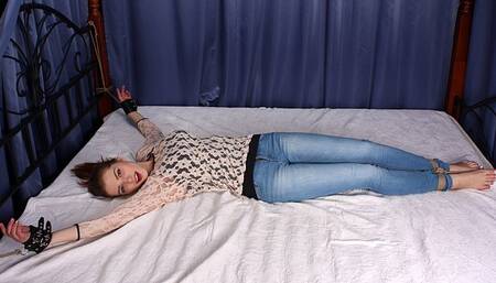 Lisa: smiling barefoot girl in top and jeans, tied on a bed with her hands locked in leather cuffs and spread wide and her ankles bound together and tied to the footboard of the bed, is wiggling on the bed (HD MP4)